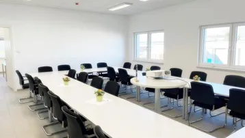 Heller Konferenzraum mit langen Tischen, vielen Stühlen und großen Fenstern für Besprechungen.