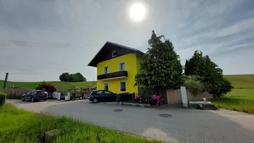 Gelbes Haus mit Balkon und dunklem Dach, umgeben von grünen Feldern unter sonnigem Himmel.