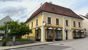 Historisches gelbes Gebäude mit rotem Ziegeldach und großen Schaufenstern an einer Straßenecke.
