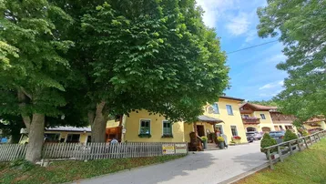 Gelbes Gebäude mit traditioneller Architektur, umgeben von üppigem Grün und Bäumen unter blauem Himmel.