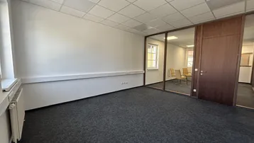 Geräumiges Büro mit Teppichboden, weißen Wänden und Glasabtrennung zu einem weiteren Raum.