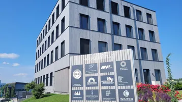Modernes Geschäftsgebäude mit Holzfassade und großem Informationsschild vor blühenden Pflanzen unter blauem Himmel.