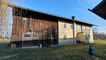 Ländliches Anwesen mit traditionellem Wohnhaus und angrenzender Holzscheune unter blauem Himmel.