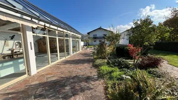 Ein sonniger Gartenbereich mit einem Wintergarten und einem gepflasterten Weg, umgeben von Grünflächen.