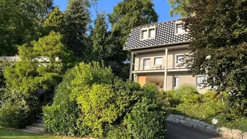 Wohnhaus mit dunklem Dach und Balkonen, umgeben von üppigem Grün und Bäumen an einem sonnigen Tag.