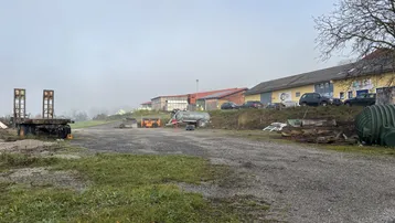 Großer Außenbereich mit landwirtschaftlichen Geräten und Gebäuden im Hintergrund unter bewölktem Himmel.