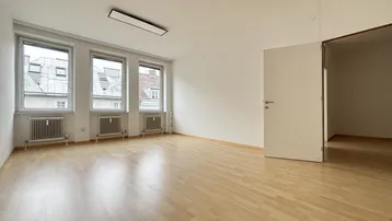 Heller, leerer Raum mit Holzparkettboden, drei Fenstern und Blick auf umliegende Gebäude.
