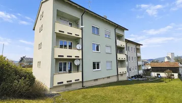 Mehrfamilienhaus mit hellgrüner Fassade, Balkonen und großem Rasenbereich unter blauem Himmel.