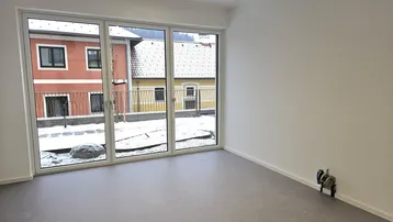 Heller, leerer Raum mit großen Fenstern, die einen Blick auf einen verschneiten Innenhof und gegenüberliegende Gebäude bieten.