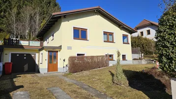 Gelbes Einfamilienhaus mit braunen Fenstern, Garage und Auffahrt, umgeben von Bäumen.