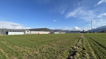 Weitläufiges grünes Feld mit Industriebauten im Hintergrund und einer malerischen Bergkulisse unter blauem Himmel.