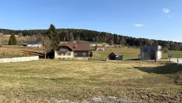 Weitläufige Grünfläche mit Blick auf bewaldete Hügel und vereinzelte Häuser in der Ferne unter blauem Himmel.