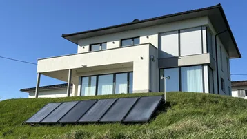 Modernes Haus mit Photovoltaikanlage auf dem grünen Hang im Vordergrund, unter blauem Himmel.