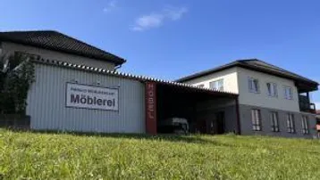 Gebäude mit einer Möblerei-Werbetafel und einem angrenzenden Wohnhaus unter blauem Himmel.