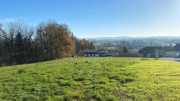 Weitläufige grüne Wiese mit Blick auf die umliegende Landschaft und entfernte Bebauung unter blauem Himmel.