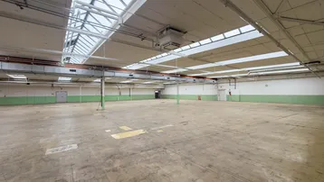 Geräumige, leere Industriehalle mit hohen Decken, Oberlichtern und Betonboden, ideal für Lagerung oder Produktion.