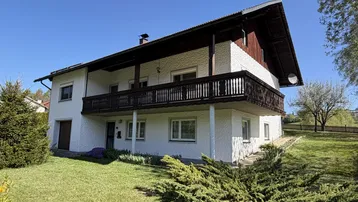 Traditionelles Haus mit weißer Fassade, Holzbalkon und gepflegtem Vorgarten unter blauem Himmel.