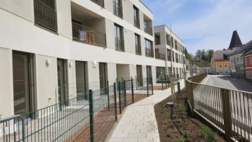 Modernes Mehrfamilienhaus mit Balkonen und Erdgeschosswohnungen, umgeben von einem gepflasterten Weg und Grünflächen.