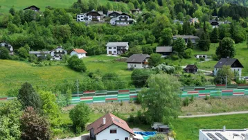 Luftaufnahme einer Wohnsiedlung in einer grünen Berglandschaft mit schneebedeckten Gipfeln und modernen Gebäuden.