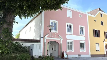 Rosa gestrichenes Gebäude mit weißen Fensterrahmen und einem großen Baum im Vordergrund, an einem sonnigen Tag.