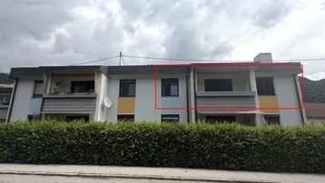 Mehrfamilienhaus mit Balkonen und einer grünen Hecke im Vordergrund unter bewölktem Himmel.