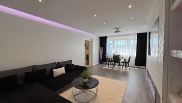 Modernes Wohnzimmer mit integriertem Essbereich, indirekter Beleuchtung und Holzboden.
