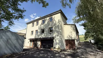 Mehrstöckiges Gebäude mit heller Fassade, umgeben von Bäumen und einer Zufahrt unter blauem Himmel.