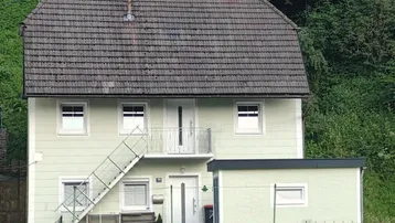 Einfamilienhaus mit hellgrüner Fassade, Satteldach und kleinem Balkon, umgeben von Bäumen und Natur.