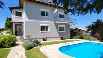Hausfassade mit Garten und Pool unter blauem Himmel an einem sonnigen Tag.