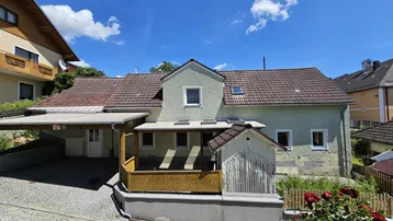 Ansicht des Wohnhauses mit Carport, Satteldach und kleiner Gartenhütte unter blauem Himmel.