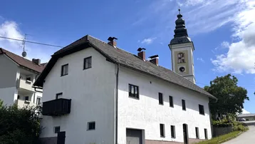 Weißes Wohnhaus mit Satteldach und kleinem Balkon, im Hintergrund ein Kirchturm unter blauem Himmel.