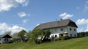 Ländliches Wohnhaus mit weißer Fassade und dunklem Dach, umgeben von grünen Wiesen unter blauem Himmel.