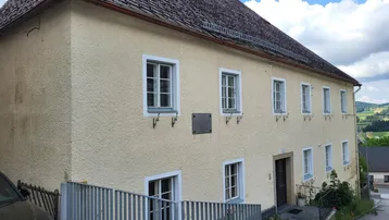 Traditionelles Gebäude mit heller Fassade, Satteldach und mehreren Fenstern in ländlicher Umgebung.