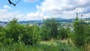 Weitläufiger Blick über grüne Hügel und eine ländliche Siedlung unter einem blauen Himmel mit Wolken.