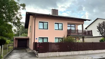Einfamilienhaus mit rosafarbener Fassade, Balkon und integrierter Garage unter bewölktem Himmel.