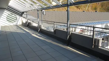 Große, geflieste Terrasse mit Glasdach und Blick auf die Umgebung und Bäume.