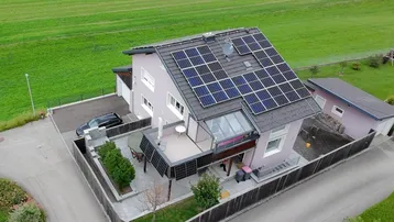 Luftaufnahme eines gepflegten Einfamilienhauses mit Solaranlage auf dem Dach, Garten und Terrasse, umgeben von grünen Feldern.