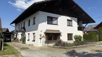 Weißes Einfamilienhaus mit traditionellem Satteldach, Carport und gepflasterter Auffahrt unter blauem Himmel.