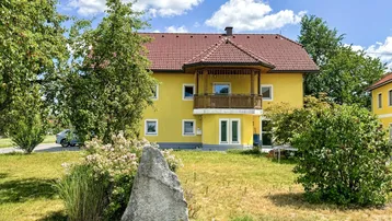 Gelbes Wohnhaus mit rotem Ziegeldach, Balkon und großem Vorgarten unter blauem Himmel.