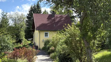 Gelbes Einfamilienhaus mit rotem Ziegeldach, umgeben von üppigem Grün und Bäumen.