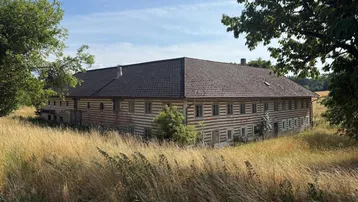 Großes ländliches Gebäude mit Holzbalken und Ziegeldach, umgeben von hohen Gräsern.