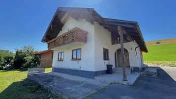 Traditionelles Haus mit Holzbalkon und Satteldach in ländlicher Umgebung unter strahlendem Sonnenschein.