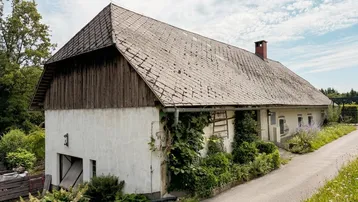 Ländliches Gebäude mit traditionellem Dach und bewachsenen Fassadenbereichen unter bewölktem Himmel.
