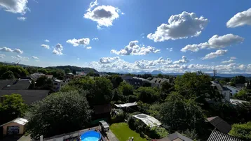 Weitläufiger Blick über die Dächer der Nachbarschaft mit viel Grün und Bergen im Hintergrund an einem sonnigen Tag.