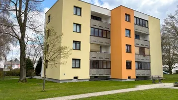 Mehrfamilienhaus mit gelber und oranger Fassade, umgeben von einer grünen Rasenfläche und Bäumen.