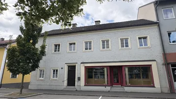 Vorderansicht eines älteren Gebäudes mit hellgrauer Fassade und großen Schaufenstern im Erdgeschoss.