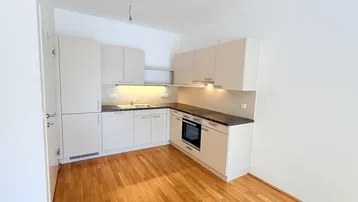 Moderne Einbauküche mit hellen Schränken, dunkler Arbeitsplatte und Holzboden in einem leeren Raum.