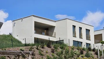 Modernes Wohnhaus mit Flachdach und integriertem Balkon, eingebettet in eine grüne Hanglage.