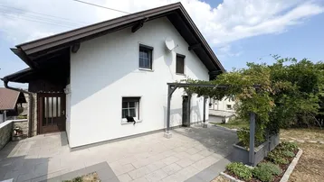 Weißes Haus mit braunem Dach und einer gepflasterten Terrasse mit Pergola im Gartenbereich.