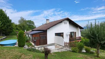 Weißes Einfamilienhaus mit braunem Dach, Garten, kleinem Pool und Balkon unter blauem Himmel.